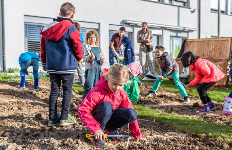 Bambini che coltivano un orto scolastico insieme a un’insegnante, in un giardino verde all’aperto